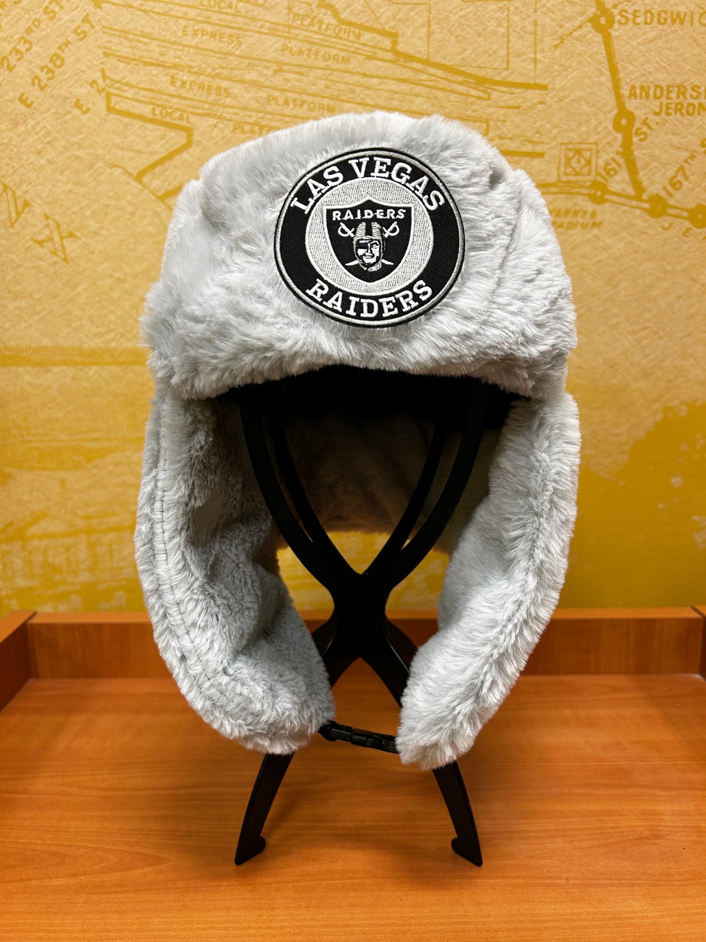 A plush light gray and white fuzzy cap with a black and white Las Vegas Raiders logo patch on the side, shown resting on a wooden surface.